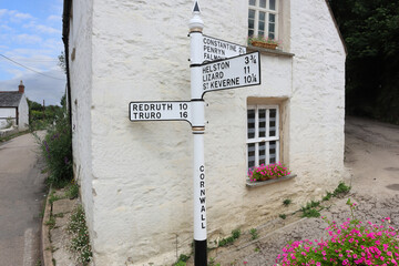 An old fashioned cast iron sign directs travellers to famous towns and villages in Cornwall, with backdrop of a typical Cornish holiday cottage