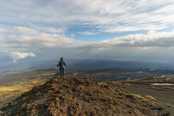 Hiking person looking at view of volcanic landscape of Mount Etna with black lava and yellow grass on partly cloudy winter day, Catania, Sicily, Italy