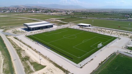 An aerial view of a modern soccer field with artificial turf. The field is surrounded by a gravel parking lot and a grassy area.