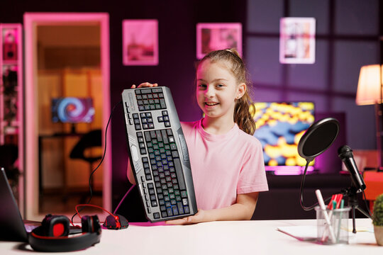 Happy charismatic girl presenting latest gaming computer peripheral tech on her internet channel. Cheerful viral internet star kid filming review of wired mechanical keyboard