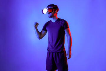 Happy man with VR goggle exercising and stretching arm at neon light background. Caucasian person with casual cloth relaxing and exercising by using virtual reality simulator technology. Deviation.