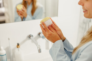 A woman elegantly holds a luxurious skincare cream in her stylish, modern bathroom, emphasizing her...
