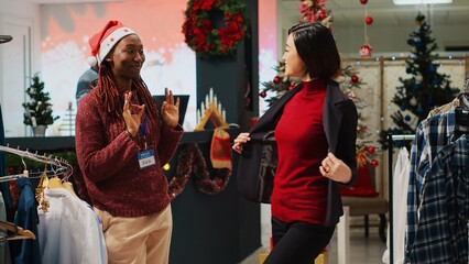 Asian businesswoman doing pirouette while trying stylish blazer, asking retail assistant for opinion before making purchase. Customer and employee in Christmas decorated clothing shop © DC Studio