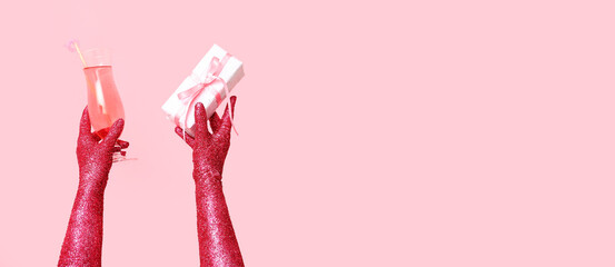 Female hands covered with glitter holding glass of cocktail and gift on pink background with space for text