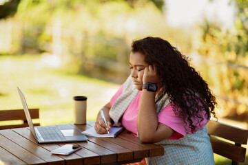 A young woman sits at a wooden table, deep in thought while writing notes in a notebook, enjoying...