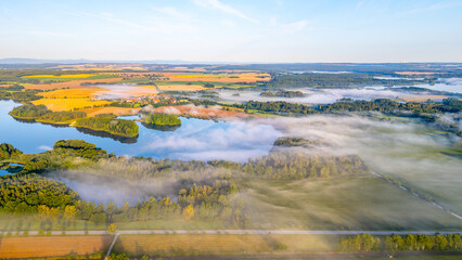 A tranquil morning unveils a hazy view of Rozmberk Pond surrounded by gently rolling hills and lush greenery. The mist hangs low over the water, creating a serene atmosphere in Trebon, Czechia.