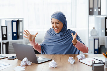 Long-awaited project success. Euphoric businesswoman looking at laptop screen and raising hands in excitement, happy after completing job