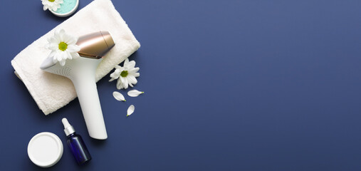 Modern photoepilator with beautiful flowers, bath towel and cosmetic products on dark blue background
