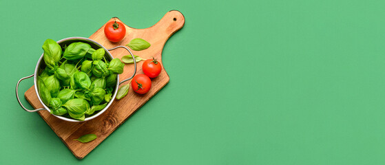 Wooden board with cooking pot of fresh green basil and tomatoes on color background