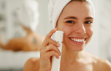 A woman is happily enjoying her skincare routine while using a beauty device and has a radiant, cheerful smile