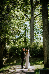 Fototapeta premium A bride and groom stand hand in hand on a tree-lined path, bathed in sunlight, surrounded by lush greenery on their wedding day.