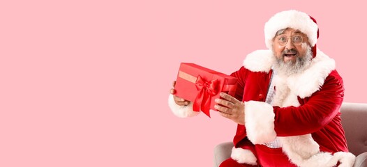 Santa Claus with Christmas present sitting in armchair on pink background with space for text