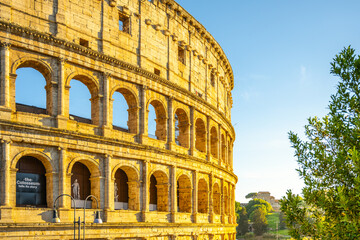 Fototapeta premium The ancient Colosseum stands majestically in Rome, illuminated by the warm glow of the evening sun. Visitors can explore its historic arches and vibrant surroundings, illustrating timeless beauty.