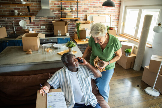 Senior couple unpacking boxes during move into new home with checklist and phone call