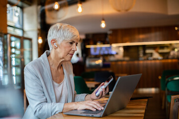 Senior businesswoman working on laptop in a cafe with wireless earbuds