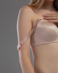 Obraz premium A woman gently holds her bra strap while trying to adjust it. She stands in a neutral gray space, highlighting the outfit and her careful motion.