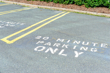 Obraz premium An asphalt parking lot with yellow double striped dividers. A white letter with 20-minute parking only painted on the ground. There's hedging and mulch as a border around the edge of the lot. 