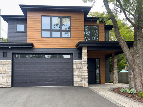 A street view of a modern suburban-style house. There's a patio, the exterior walls are faux wood siding and beige rock, and the garage door is a large brown metal door with small glass windows.   