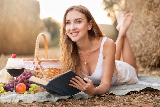 Beautiful woman in white blouse and shorts lying down and and reading book near haystacks with basket of fruit and red wine, fashion concept