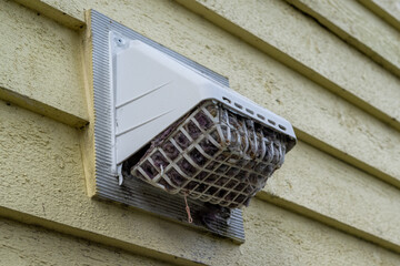 The exterior of a bright yellow house wall with a white dryer vent exposes the air vent which is...