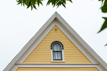 Obraz premium The exterior wall of a bright yellow country cottage with white trim. The historic building has an arched shaped double hung window, with black trim, in the center of the top floor of the pitched roof