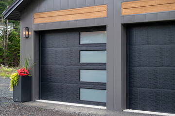 Fototapeta premium A modern black panel metal exterior garage doors with four small horizontal frosted glass windows. The modern door is on a luxury dark grey contemporary house with a concrete driveway and flower pot. 