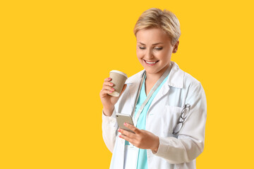 Female medical intern with cup of coffee using mobile phone on yellow background