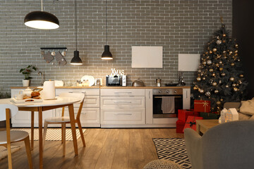 Interior of modern kitchen with Christmas tree, glowing lamps and dining table at evening