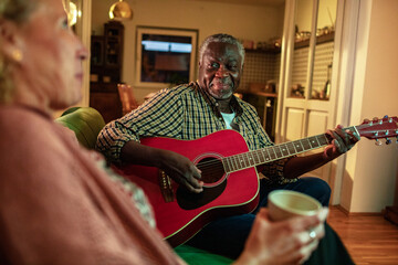 Joyful senior man playing guitar for his wife at home during evening