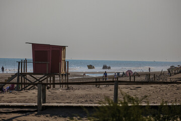 Photo of a casual day on the beaches of the Atlantic Ocean, featuring a wooden lifeguard house, an umbrella, and an artisanal fishing boat.