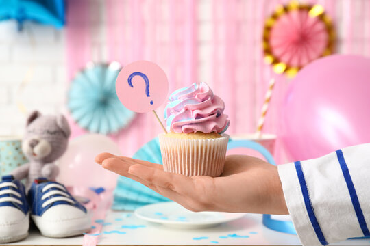 Female hand with delicious cupcake, baby booties and decorations on table against white brick background. Gender reveal party concept