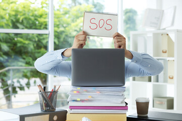 Man holding notebook with word SOS and hiding face behind laptop in office