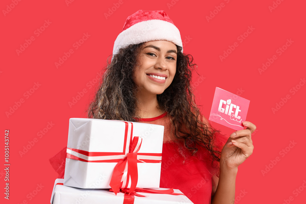 Sticker Beautiful young happy African-American woman in Santa hat with present boxes and gift card on red background. Christmas shopping concept