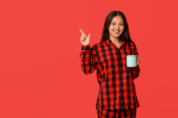 Beautiful young Asian woman in checkered pajamas with cup of coffee pointing at something on red...