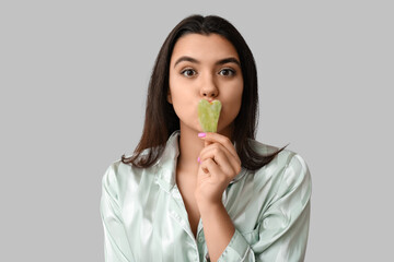 Beautiful young woman with guasha facial tool on grey background