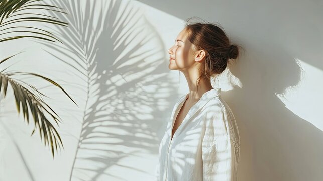 A woman stands with her eyes closed, bathed in the soft glow of sunlight streaming through a window, with the shadow of a palm tree cast on the wall behind her.