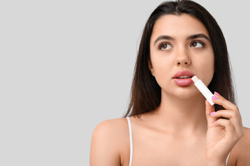 Young woman applying lip balm on grey background