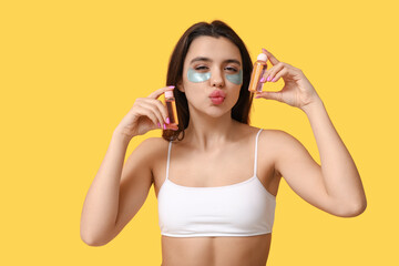 Beautiful young woman with under-eye patches holding bottles of cosmetic products on yellow background