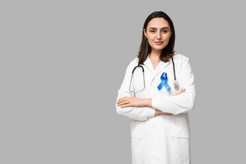 Young female doctor with blue ribbon on grey background. Colon cancer awareness concept