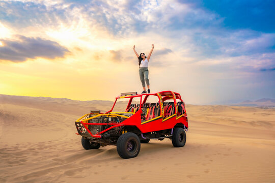 Young woman celebrating on top of a colorful dune buggy in the Huacachina desert, Ica, Peru, enjoying the sunset and adventure.