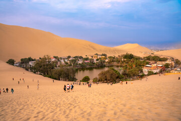 A stunning view of the Huacachina oasis surrounded by towering sand dunes, located in Ica, Peru, at dusk.