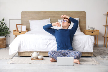 Morning of handsome young man in stylish pajamas with teapot and laptop sitting at home