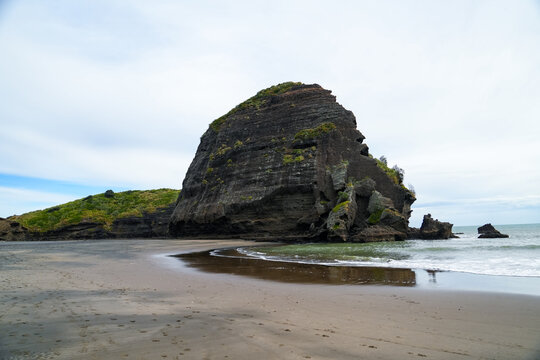 A majestic rocky outcrop dominates the sandy beach in this coastal scene. The calm waves wash gently against the shore, and the massive rock formation creates a striking contrast with the expansive sk