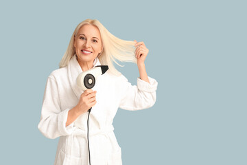 Fototapeta premium Mature woman in bathrobe with hair dryer on color background