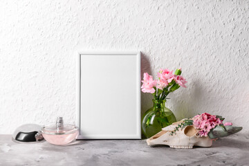 Beautiful blank photo frame, bottle of perfume and skull with flowers on grey table near white wall