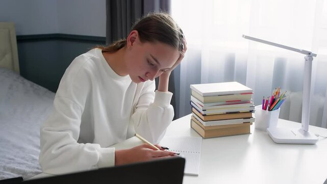 Teenage girl is sitting at her desk, struggling to concentrate on her homework with her head in her hand. She is surrounded by books and school supplies. Track in shot. Zoom in