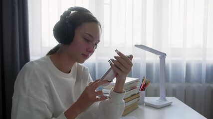 Teenage girl is sitting at a desk with books and school supplies, wearing headphones and using a smartphone while doing her homework