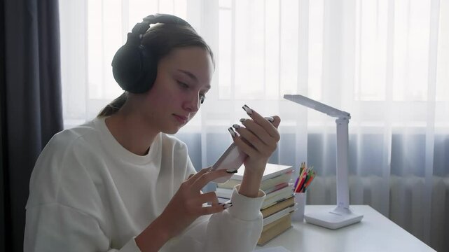 Teenage girl is sitting at a desk with books and school supplies, wearing headphones and using a smartphone while doing her homework