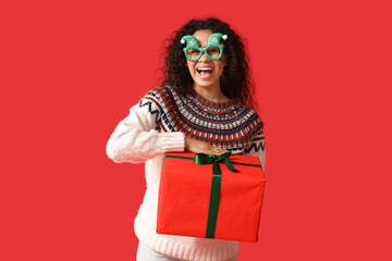 Happy young African-American woman in Christmas sweater and funny glasses with gift box on red background