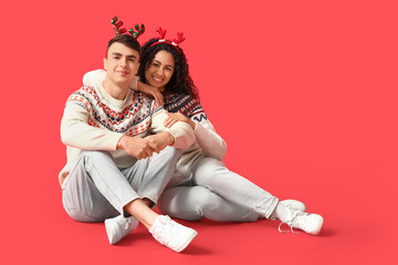 Happy young couple in Christmas sweaters sitting on red background